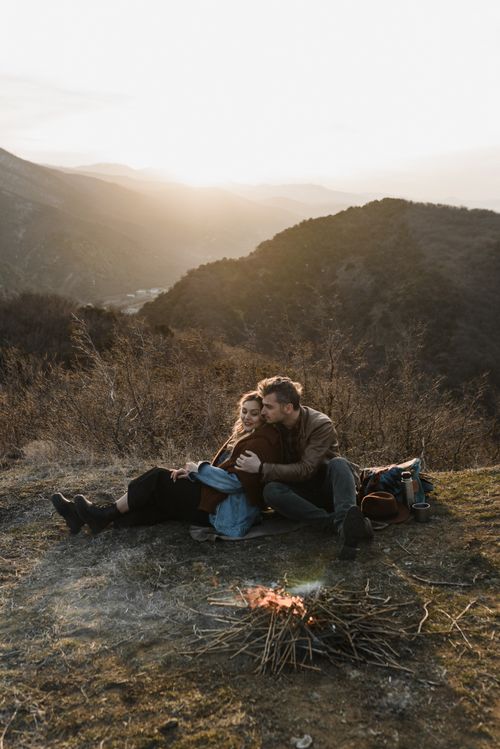 Free A couple embraces near a small fire in a mountainous landscape at sunset, symbolizing love and adventure. Stock Photo