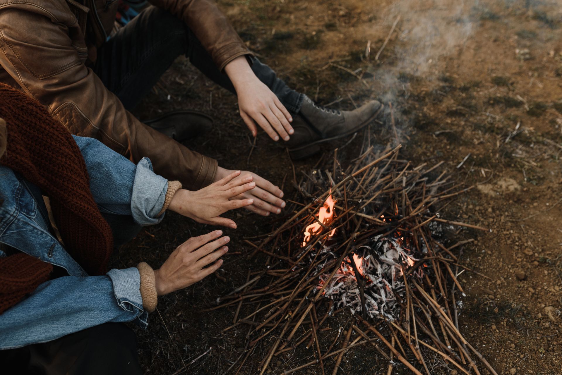 Two individuals warming their hands by a small campfire made of sticks, creating a cozy outdoor atmosphere.