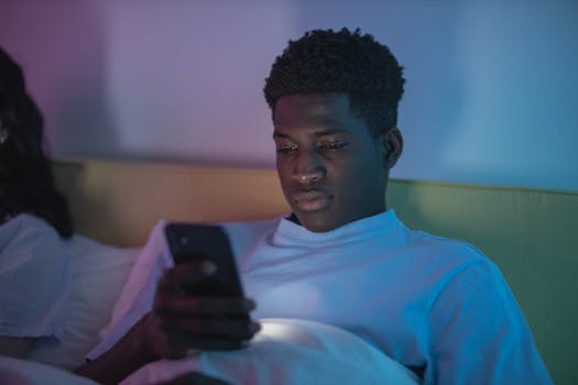 A young man in a white shirt uses a smartphone while lying in a dimly lit room.