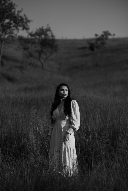 Black and white full body of peaceful ethnic female wearing long dress looking away while standing in grassy meadow in countryside