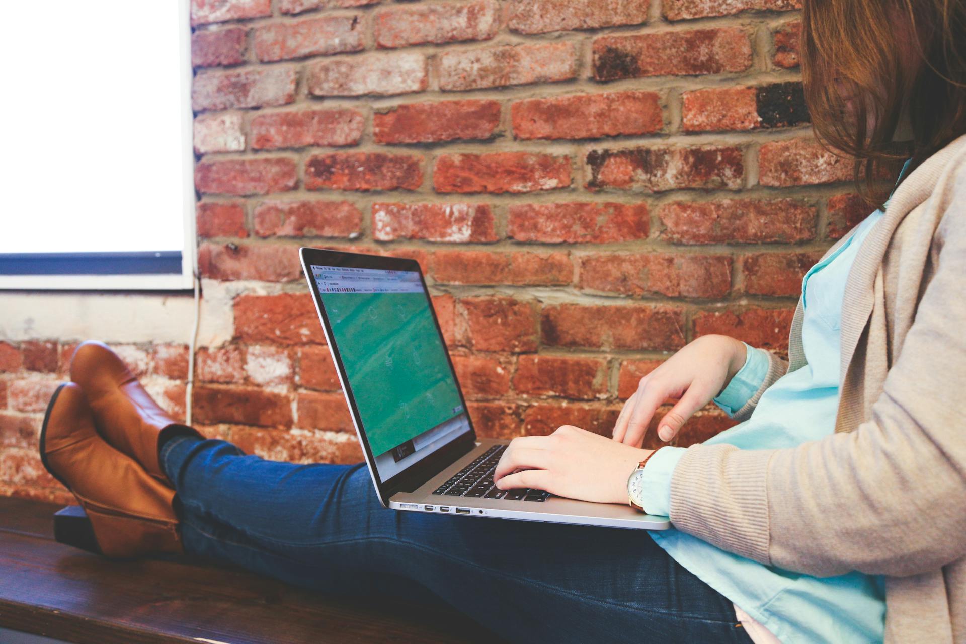 Woman working remotely on a laptop against a brick wall, representing modern flexible workspace.