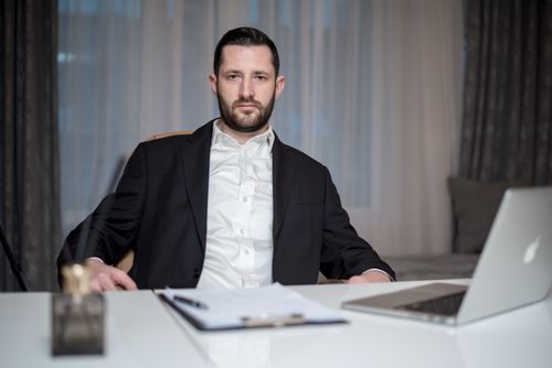 Free A serious bearded man in a black suit sits behind a desk with a laptop and documents. Stock Photo