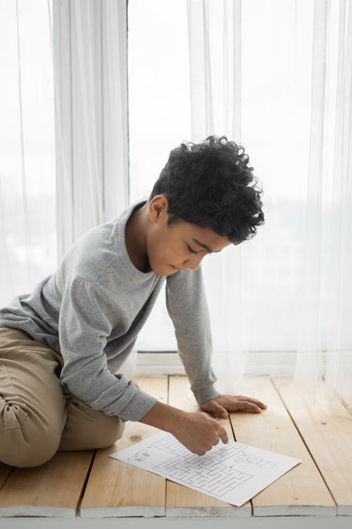 Side view of focused ethnic little boy in casual clothes sitting on wooden floor near window and trying to find correct path to solve labyrinth printed on paper