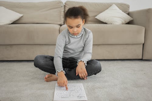 Full length of barefooted focused little black girl in casual clothes sitting on floor near sofa and solving labyrinth exercise painted on paper