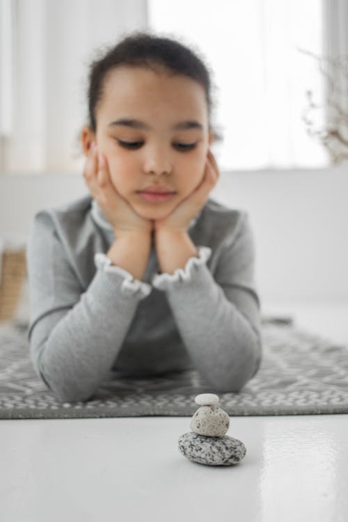 Thoughtful little African American girl in casual clothes leaning in hands while lying on floor and looking at balancing stacked stones placed at home