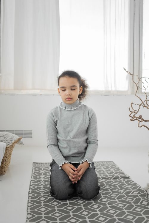Free Full body of peaceful little ethnic girl with dark Afro hair sitting on knees on carpet with closed eyes and relaxing Stock Photo