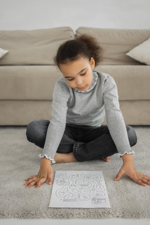 Full body of attentive little ethnic child with dark Afro hair in casual clothes solving maze task while sitting on soft carpet at home