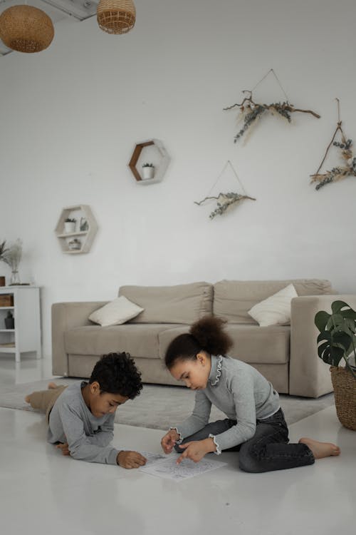 Full body of attentive little African American sister and brother in casual clothes sitting on floor and playing logical labyrinth game during weekend at home