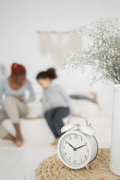 Free Unrecognizable barefooted African American mother with little daughter sitting on comfortable bed in light room near vintage alarm clock placed on table near flowers vase Stock Photo