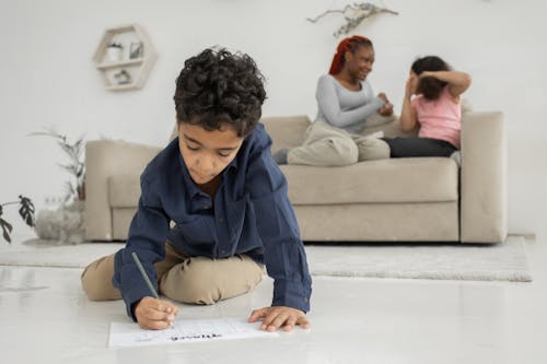 Focused little black boy doing homework on floor near mother and sister