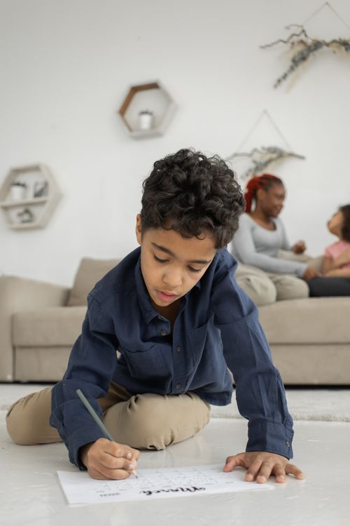 Free Concentrated African American boy with pencil writing on paper while sitting on floor in room near mother and sister on couch Stock Photo