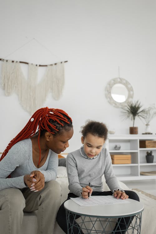 Concentrated African American mother with daughter sitting at table with paper while doing assignment together in living room at home