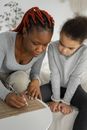 African American female in casual clothes sitting at table with adorable daughter and writing on paper with pencil