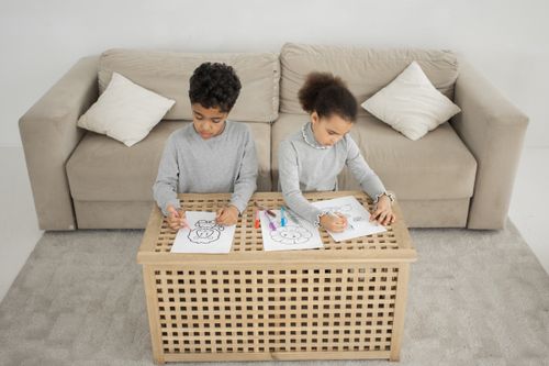 Free From above of adorable little African American children sitting at table near sofa while coloring pictures together in light living room Stock Photo