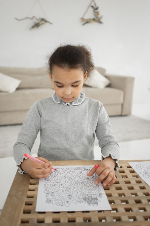 Free Concentrated little African American girl looking at paper with labyrinth while holding pen and sitting at table in light living room Stock Photo