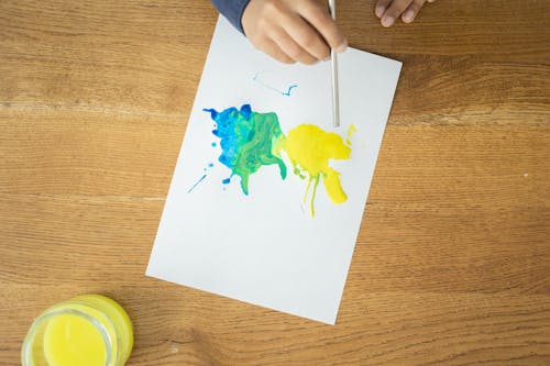Free From above of crop unrecognizable kid drawing with watercolors using straw on wooden table with glass jar of water Stock Photo