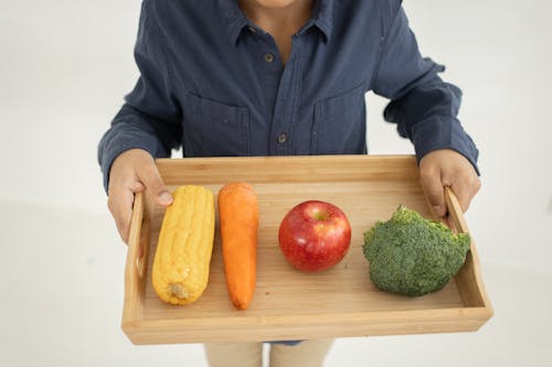 From above of crop anonymous kid in casual outfit holding wooden tray with fresh healthy broccoli apple carrot and corn against white background