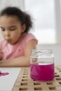 Concentrated adorable little African American girl with curly dark hair painting with straw using purple watercolor on paper while sitting at table in light room