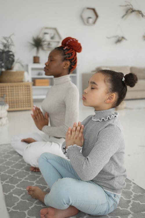 Free Side view of adorable little African American girl in casual clothes meditating with closed eyes with mother sitting on floor in Lotus pose Stock Photo