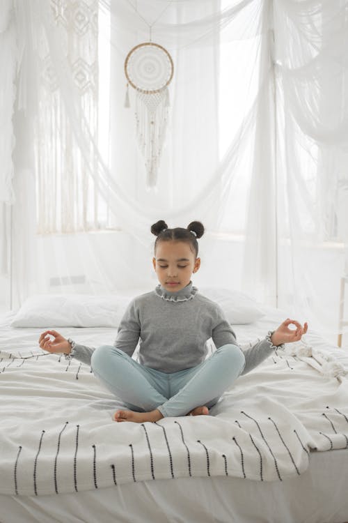 Free Full body of adorable little ethnic girl with dark hair in casual clothes sitting on comfortable bed in Lotus pose and meditating with closed eyes Stock Photo