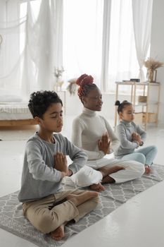 Full body of happy young black mother with son and daughter sitting on rug in Lotus pose and meditating together with closed eyes and prayer hands