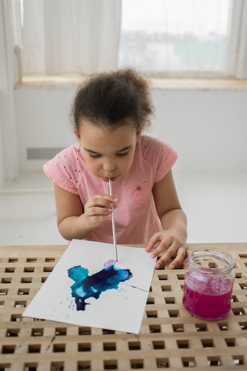 Concentrated black kid blowing watercolor with straw on paper during painting at home