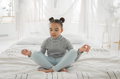 Full body of adorable little ethnic girl with pig buns in casual clothes sitting on soft bed with crossed legs while meditating with closed eyes in light bedroom