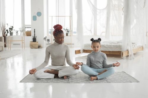 Free Full body of happy young African American woman in casual clothes sitting on floor in Lotus pose and looking at cute little daughter meditating with closed eyes Stock Photo
