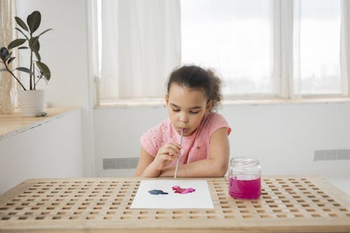 Cute little African American child with dark curly hair blowing watercolor paints on white paper while creating artwork with straw sitting at wooden table at home