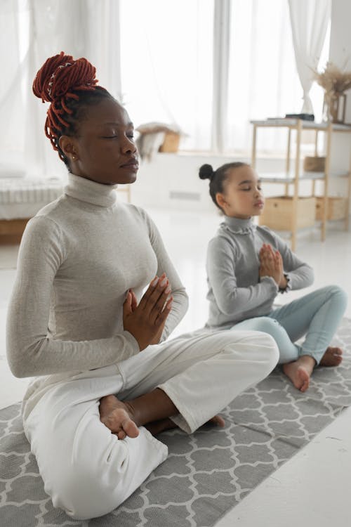 Free Full body of tranquil young barefooted African American woman in casual clothes sitting on carpet in Padmasana pose with closed eyes while meditating with adorable little daughter at home Stock Photo