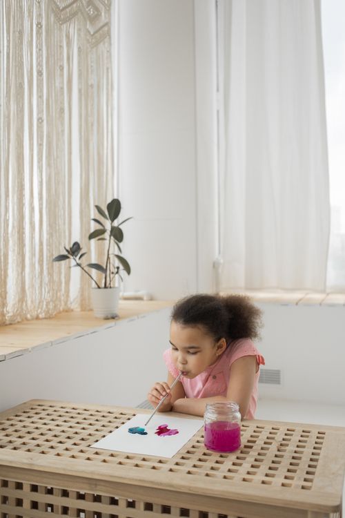 Free Cute little African American girl with curly dark hair blow painting on white paper with colorful aquarelle and straw sitting at table near window in light room Stock Photo