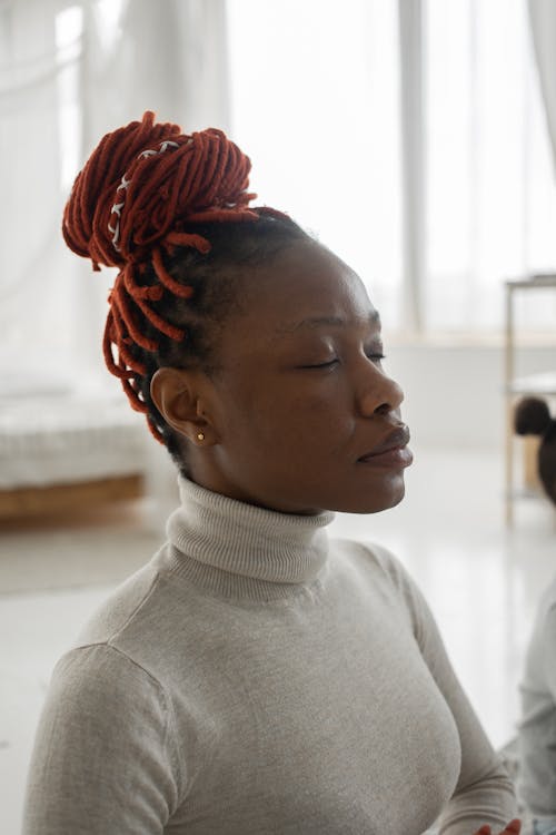 Peaceful young African American female in turtleneck with dreadlocks meditating with closed eyes in light bedroom at home