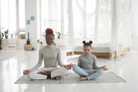 Full body of concentrated young barefooted black woman with cute little daughter sitting together on floor in Padmasana pose and mudra hands and meditating with closed eyes in light bedroom