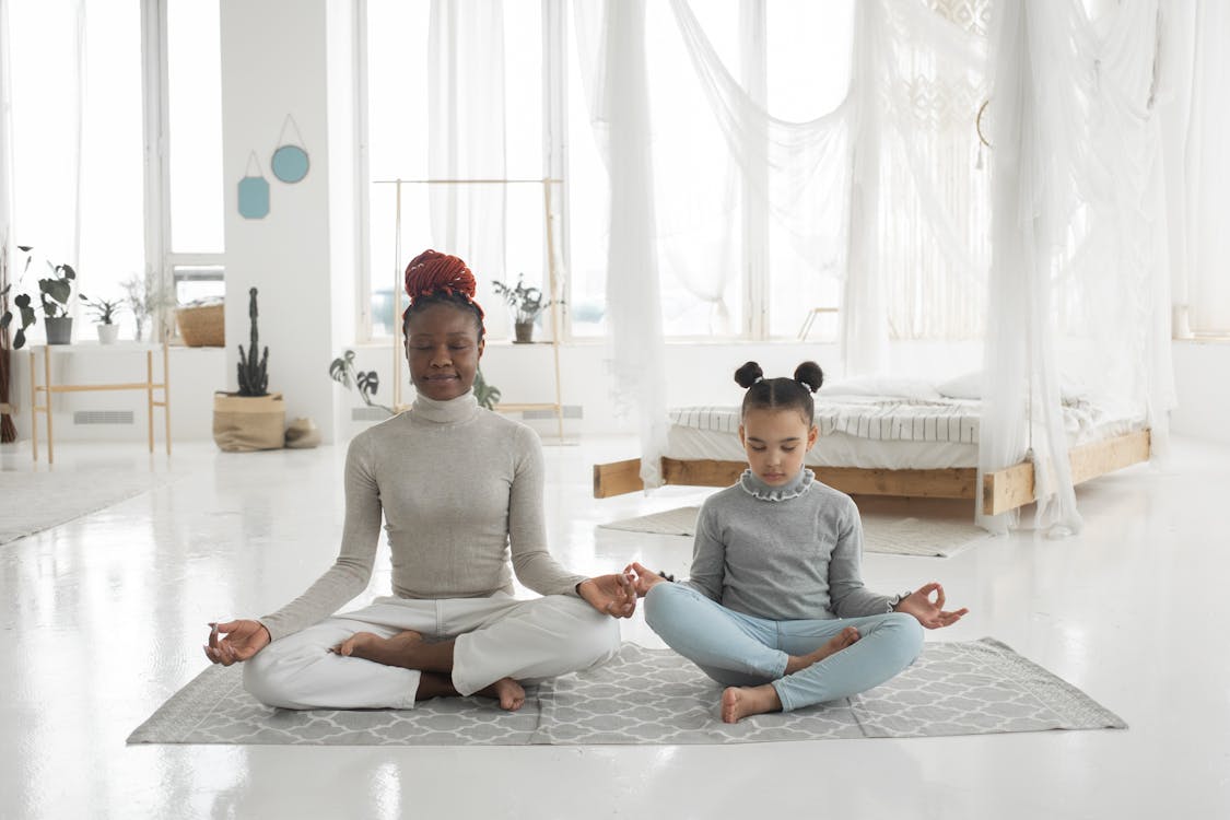 Free Full body of concentrated young barefooted black woman with cute little daughter sitting together on floor in Padmasana pose and mudra hands and meditating with closed eyes in light bedroom Stock Photo