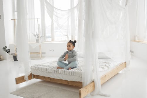 Free Full length of peaceful barefooted little ethnic kid in casual clothes meditating with closed eyes sitting on comfortable bed in Lotus pose in daylight Stock Photo