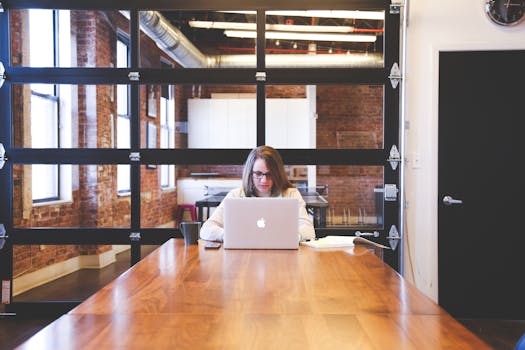 A young woman works on a laptop in a modern, industrial-style office with exposed brick walls.