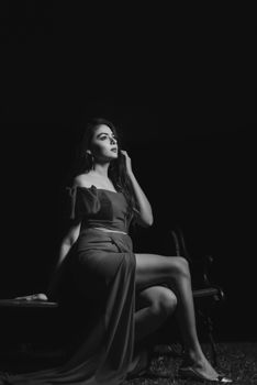 A stylish woman in a dress sits elegantly on a bench in a dramatic black and white portrait.