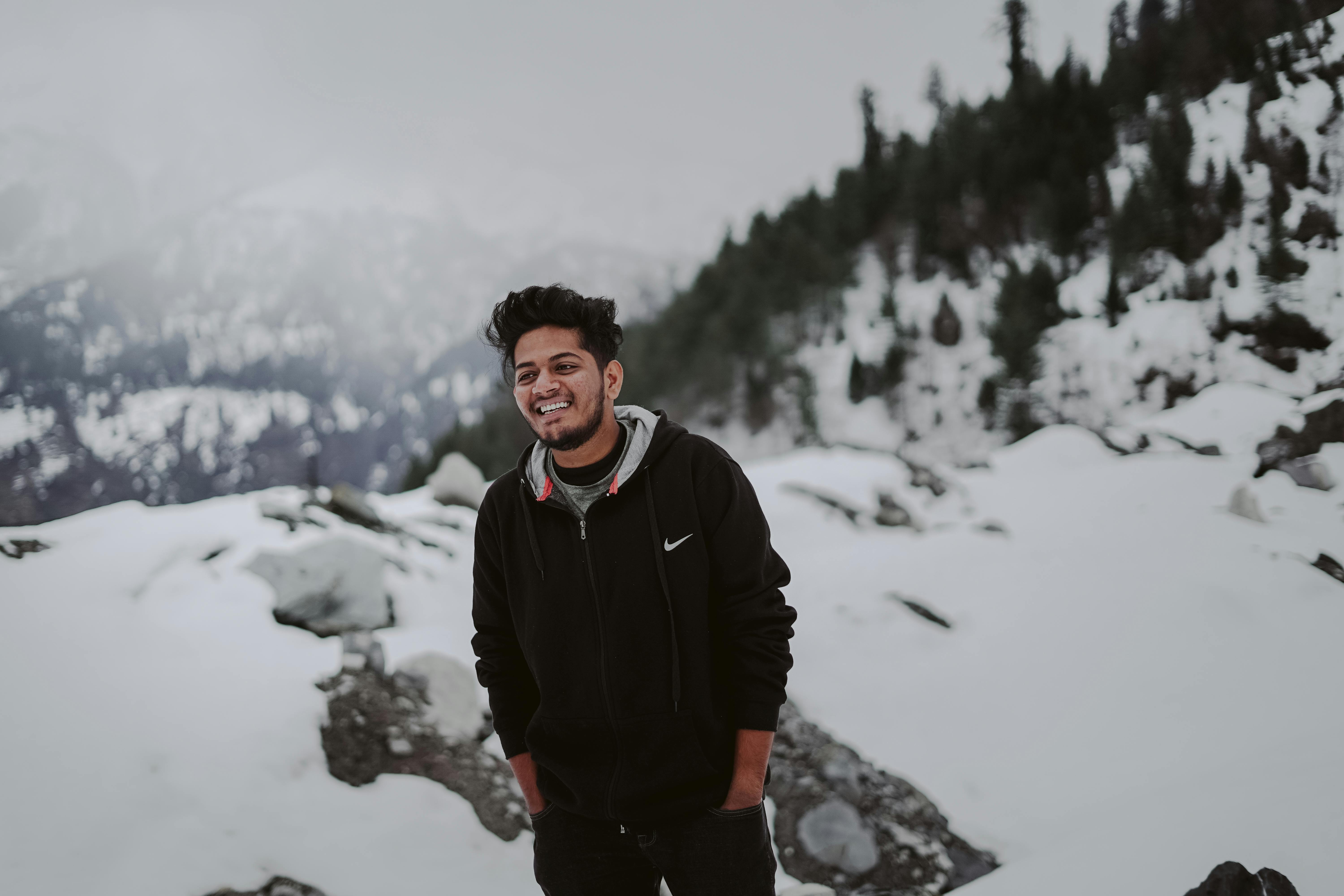 Prescription Goggle Inserts - A young man in a black jacket smiles with hands in pockets against a snowy mountain backdrop.