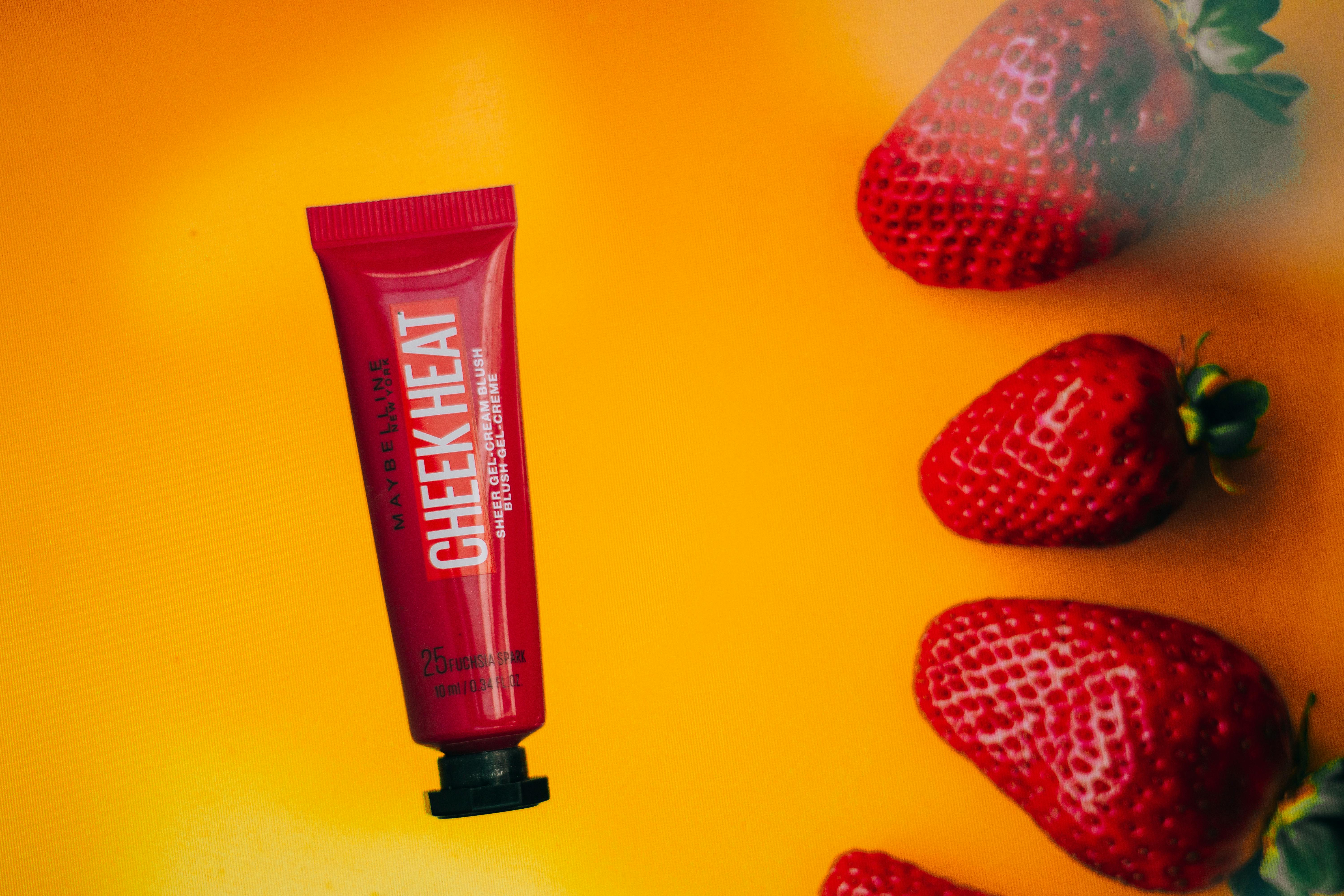 Close-up of Cheek Heat gel blush tube with strawberries on a vibrant yellow background.