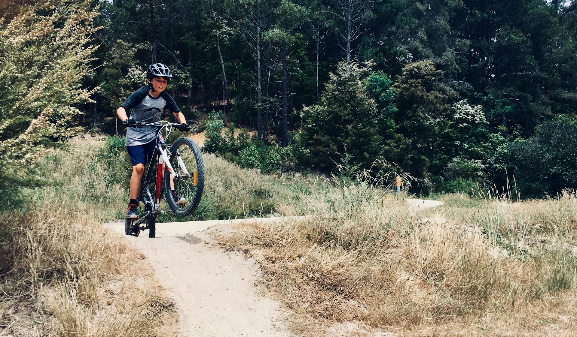 A child wearing a helmet performs a wheelie on a mountain bike in a rural forest setting.