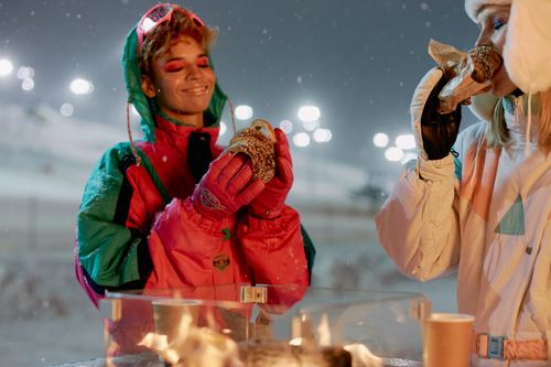Free Two women enjoy hot pastries near a fire after snowboarding at night. Stock Photo