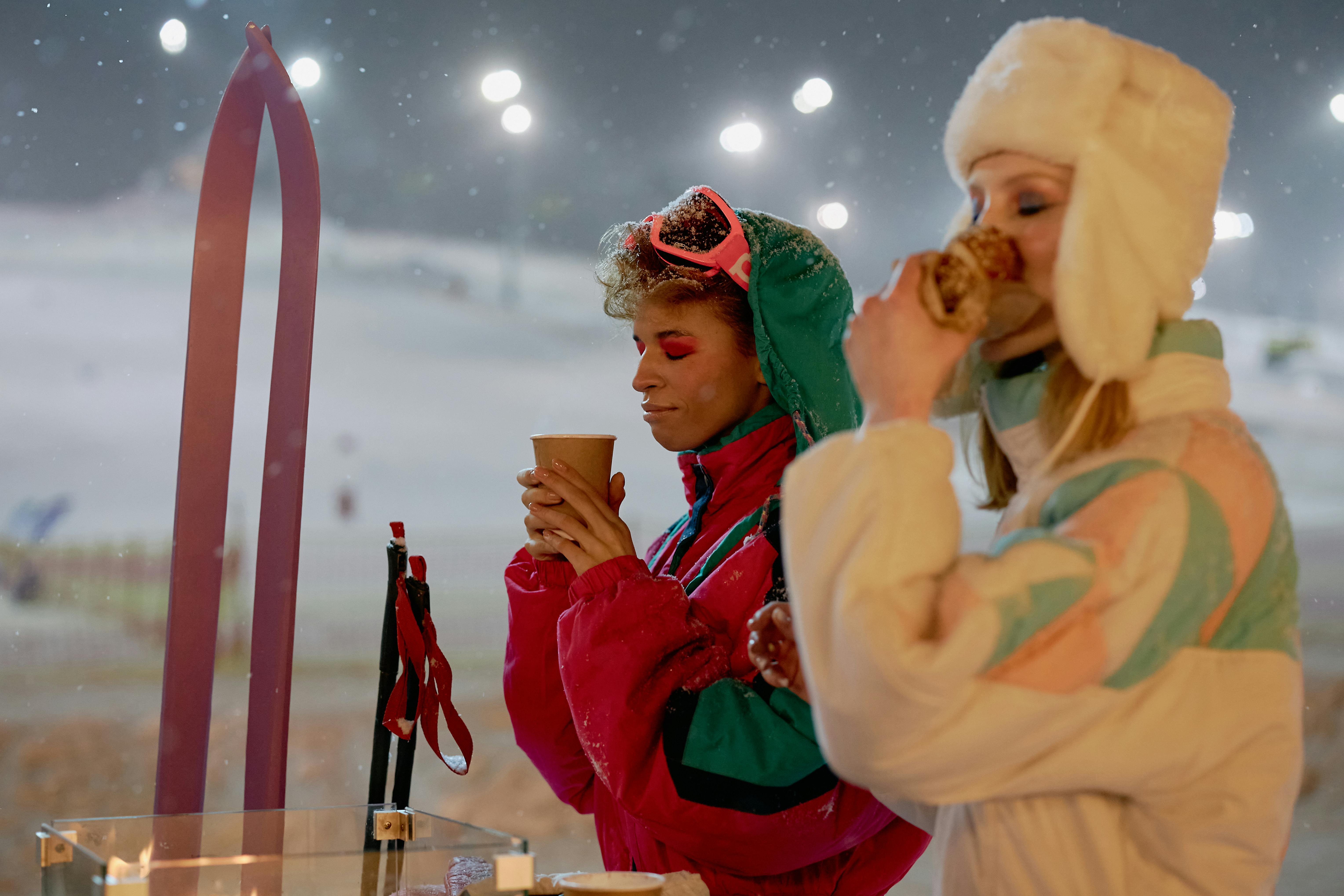 Two women enjoying hot drinks in winter ski resort, dressed warmly with skis nearby.