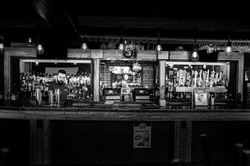 Free A black and white image of a bartender standing at a rustic bar counter in a cozy pub setting. Stock Photo