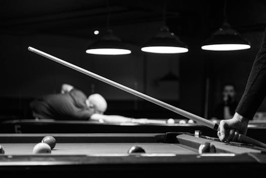 Black and white image of people playing billiards indoors, focusing on cue stick action.
