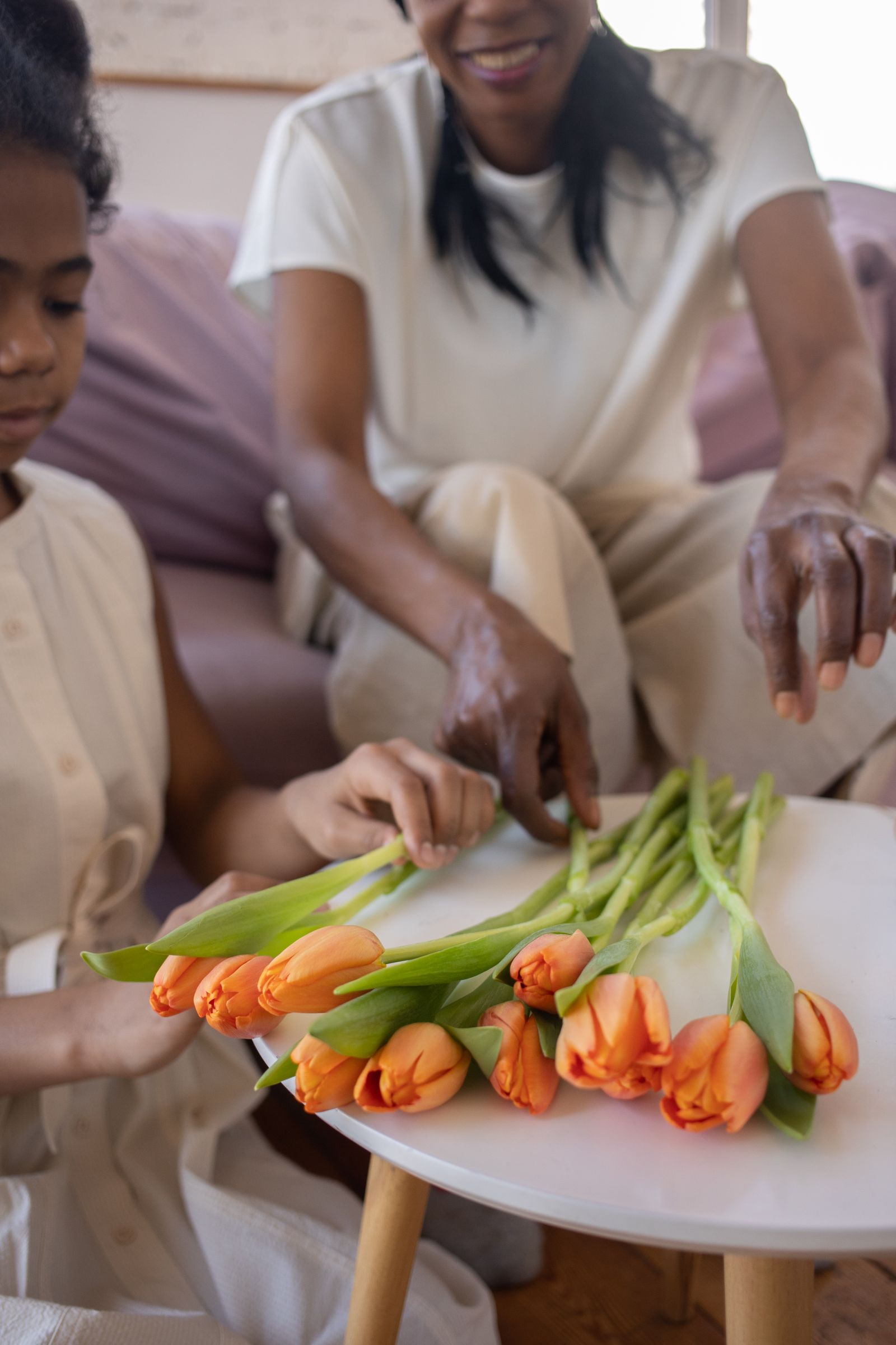 A smiling mother and her young daughter arranging bright orange tulips together on a round white table in a sunlit living room