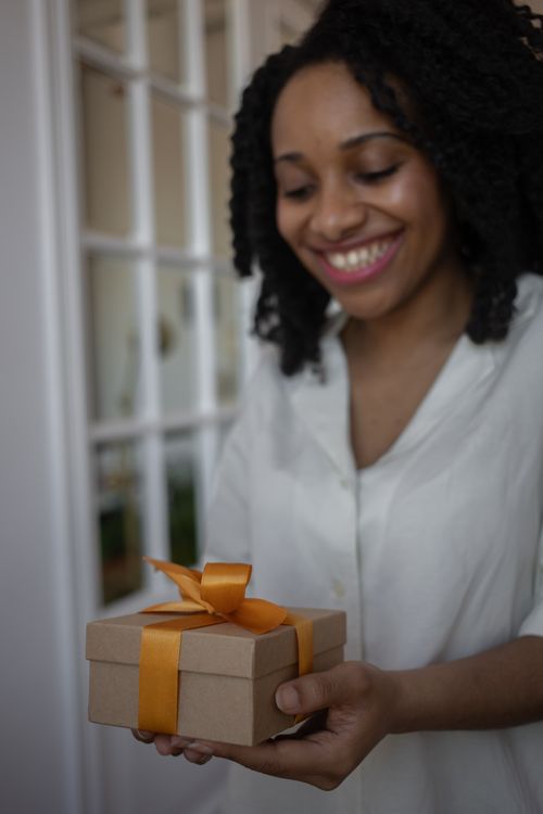 Free An African American woman joyfully holds a gift box wrapped with an orange ribbon indoors. Stock Photo