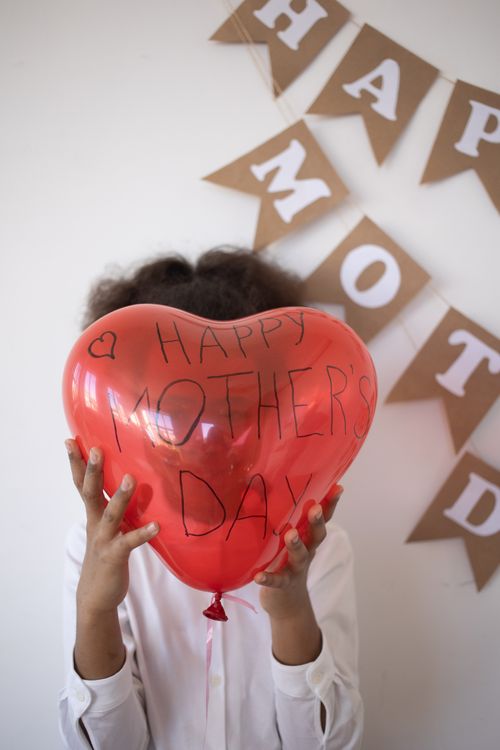 Free A child holds a heart-shaped Mother's Day balloon in front of a festive background. Stock Photo