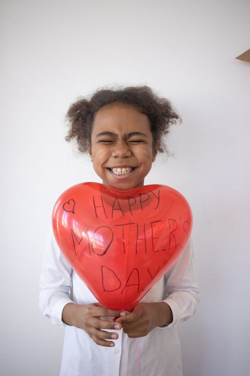 Free Joyful child holding a red heart-shaped balloon with 'Happy Mother's Day' written on it. Stock Photo