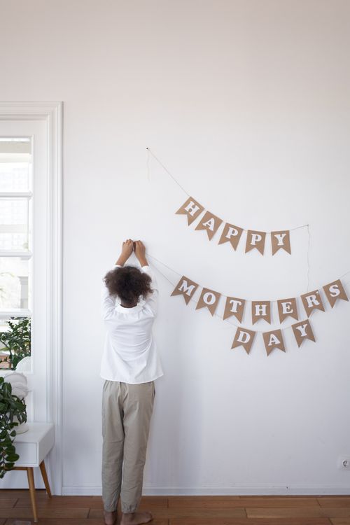 Free A child hangs a 'Happy Mothers Day' banner against a white wall indoors. Stock Photo