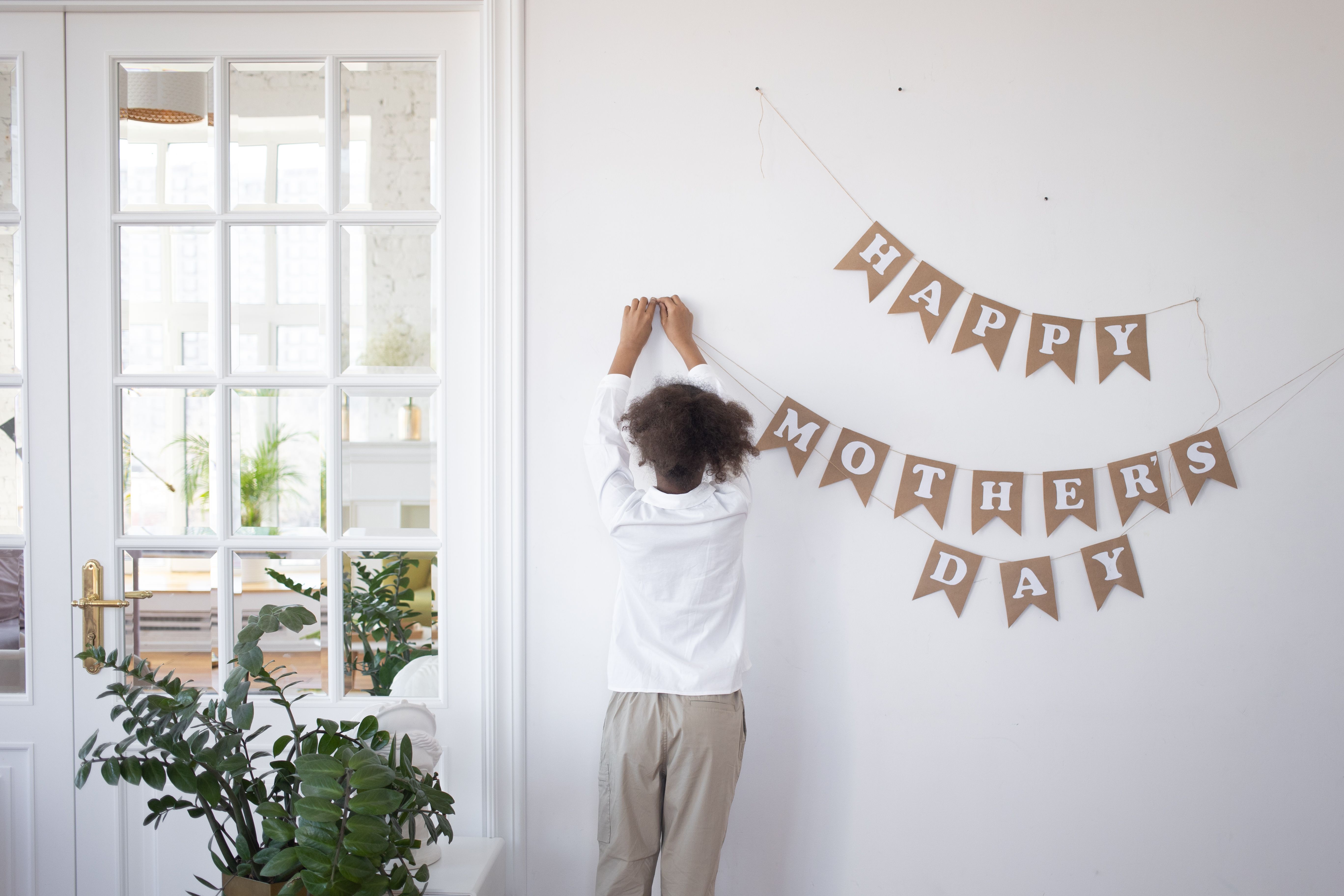A child decorates a white wall with Mother's Day banners indoors. A child decorates a white wall with Mother's Day banners indoors.